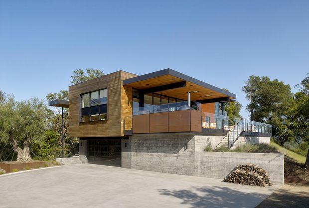 Exterior view of a contemporary multi-level house set among trees, with warm timber-clad upper walls, a broad flat roof and deep covered terrace, extensive glazing, glass balustrades, and a grey concrete-block lower level. A driveway and external stairway lead up to the elevated living areas.