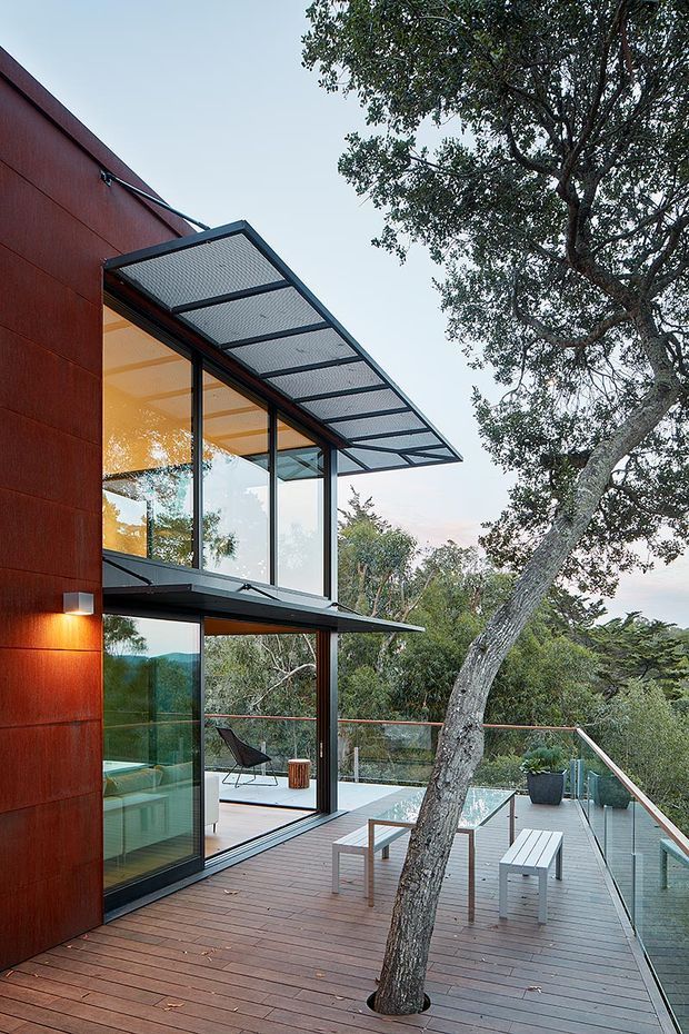 Exterior view of a contemporary multi-level home set among mature trees, with dark rust-red cladding, extensive floor-to-ceiling glazing, a projecting translucent covered balcony or awning, and a broad timber deck with glass balustrades. Outdoor seating and a small table are arranged around a tree growing through the deck, while the glazed interior is visible behind.
