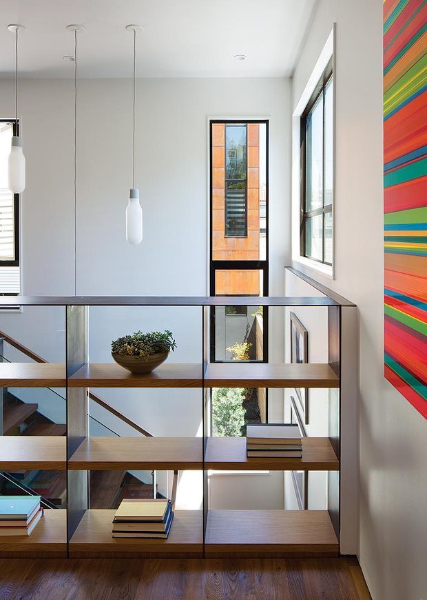 Bright contemporary stair landing with a built-in open shelving balustrade dividing the stairwell. The shelving combines dark metal framing with warm timber shelves and displays books and a small planter. Large windows and a narrow glazed door provide abundant daylight, while three slim white pendant lights hang from the high ceiling. A vivid multicoloured striped artwork forms a strong accent on the right wall. Timber flooring and visible stair elements complete the space.