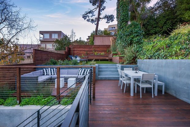 A landscaped multi-level outdoor deck and terrace with dark timber decking, built-in outdoor seating, a dining table, stairs, metal cable railings, and lush planting. The elevated urban residential setting is enclosed by timber privacy fencing and concrete retaining walls.