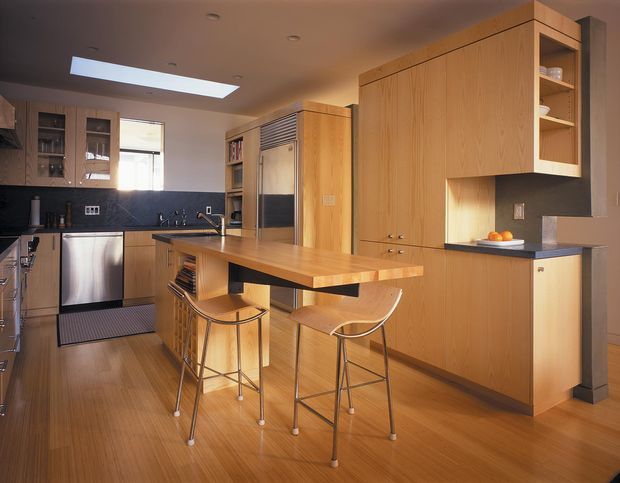 A compact, warm-toned contemporary kitchen with extensive light timber cabinetry, a central island with an integrated dining overhang and two stools, dark stone-look splashback and benchtops, stainless-steel appliances, and a skylight. Built-in storage includes open shelving and a concealed appliance or pantry zone. The space is efficiently planned and softly lit with a restrained natural material palette.