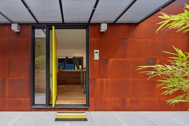 A contemporary residential entrance viewed from outside, with a bright yellow pivot door set within a dark metal-framed glazed surround. The facade is clad in rust-toned weathered steel panels, topped by a translucent or perforated covered entry canopy with black framing. A concrete path and striped entry mat lead to the doorway, while bamboo-like planting partially frames the right edge. The open door reveals a timber-floored interior and built-in timber cabinetry beyond.