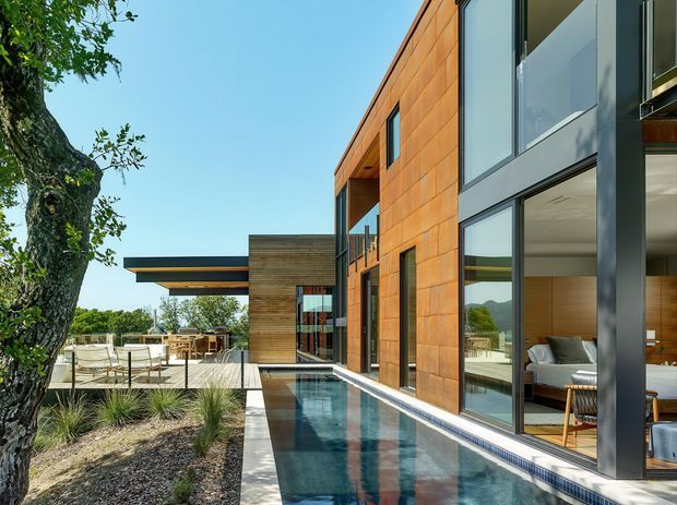 A contemporary multi-level residence viewed from its landscaped courtyard, with a long narrow reflecting pool running alongside the house. The facade combines warm orange-brown timber or timber-look cladding, dark metal window and door frames, extensive floor-to-ceiling glazing, and a projecting flat roof. Outdoor dining and lounge areas sit beneath a covered terrace, while a bright living room is visible through the glazing. Mature planting and a large tree frame the scene.