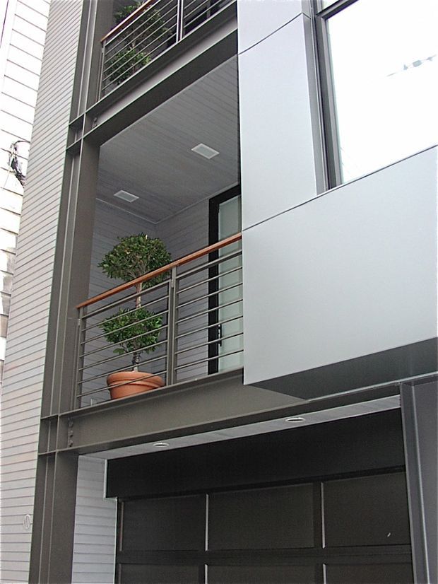 Close-up exterior view of a contemporary multi-storey building facade, featuring projecting balconies with horizontal metal railings, timber-toned handrails, dark structural framing, pale panel cladding, and large windows. A potted tree is visible on the upper balcony.