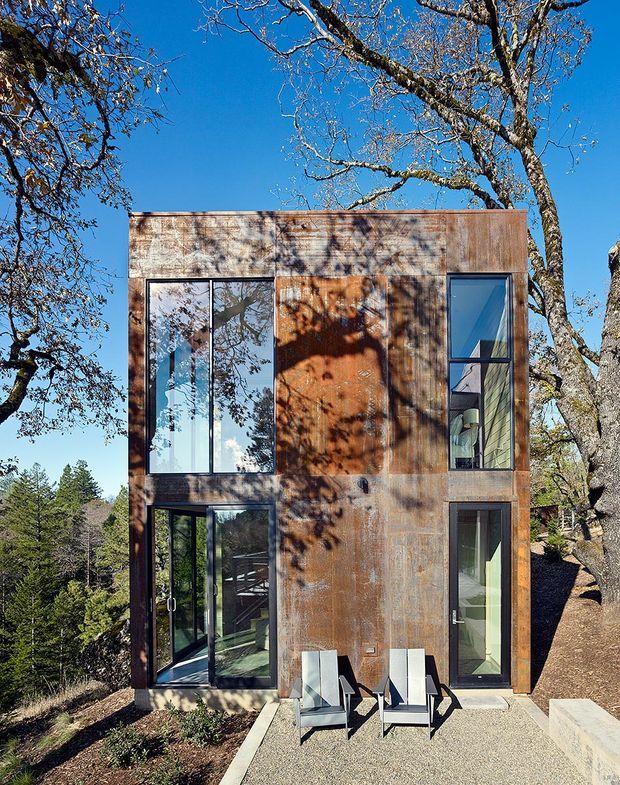 Two-storey compact contemporary dwelling set among mature trees, photographed from the exterior. The rectilinear facade is clad in weathered rust-toned metal panels, with large dark-framed windows and glazed doors. A small paved terrace with two chairs sits at the entrance, surrounded by a wooded, sloped landscape.