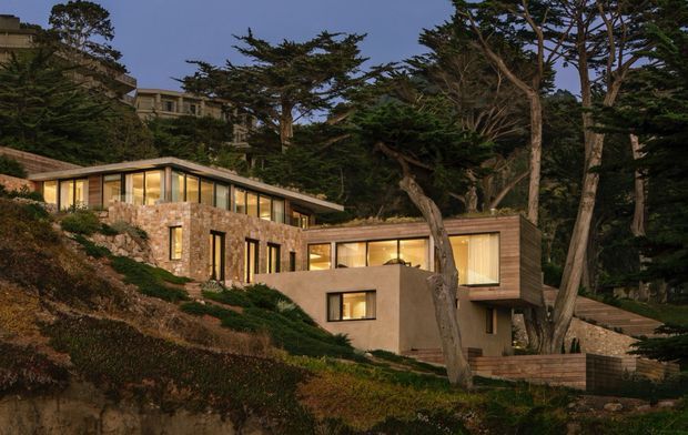 Twilight exterior view of a contemporary, multi-level residence integrated into a steep, wooded coastal-looking site. The home features extensive floor-to-ceiling glazing, warm timber-clad upper volumes, natural stone walls, earth-toned rendered surfaces, landscaped terraces, and broad low-pitched rooflines. Dense mature trees surround the property, with additional buildings visible high on the slope in the background.