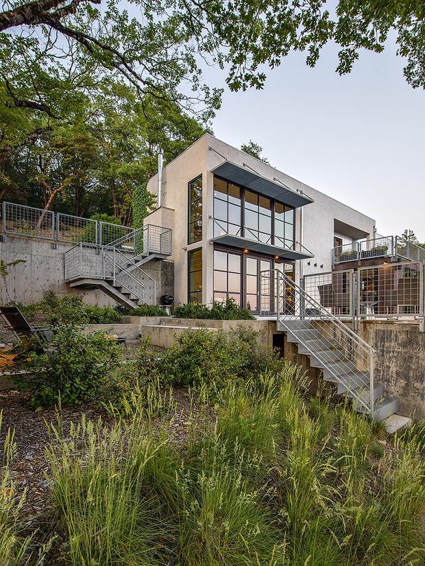 Exterior view of a contemporary multi-level residence set into a wooded, sloping site. The house has light rendered walls, extensive black-framed floor-to-ceiling glazing, projecting metal awnings, exposed concrete retaining walls and terraces, and multiple galvanized steel stairways and balustrades. Native-style grasses and dense planting surround the building.