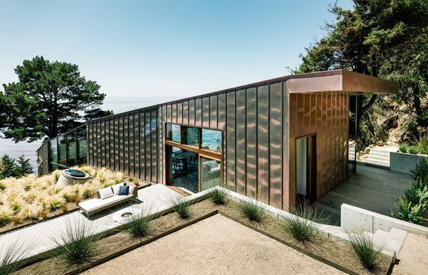 A low, contemporary pavilion-like residence set into a wooded coastal hillside. The building has a long glazed facade with dark vertical metal framing, weathered copper-toned cladding, a flat roof, and timber-lined recessed entry volume. Terraced pale paving and gravel landscaping surround an outdoor seating area, with ocean visible beyond the trees.