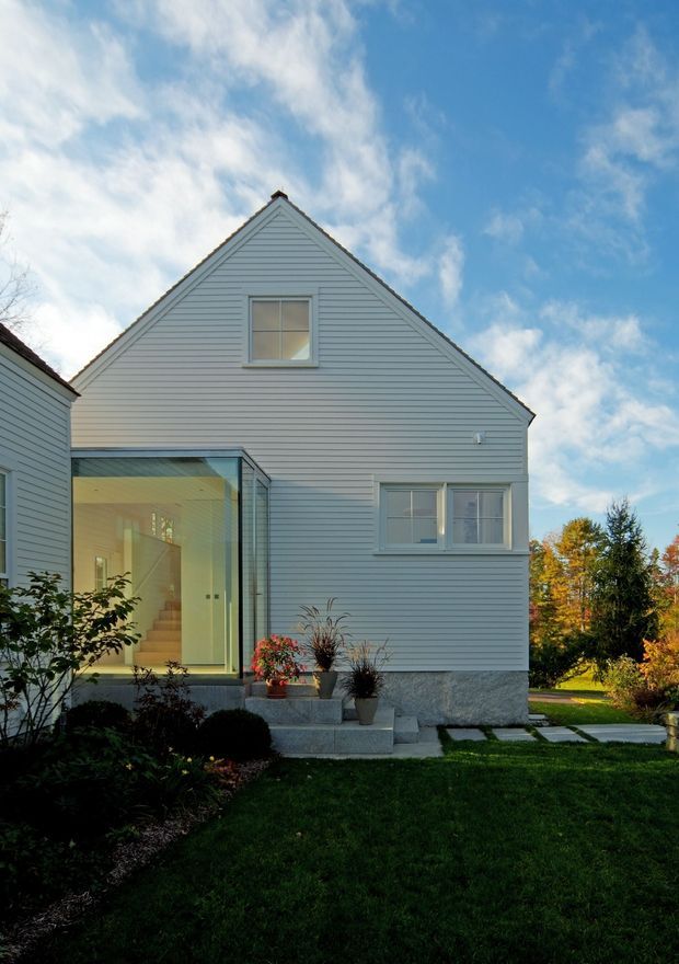 Exterior view of a compact two-storey gable-roof house clad in pale horizontal weatherboards. The facade includes a central upper window, lower multi-pane windows, and a glazed side entry or extension revealing an interior stairway. Concrete steps, potted plants, a paved stepping-stone path, lawn, and landscaped garden surround the home. The image is photographed in daylight under a partly cloudy sky.