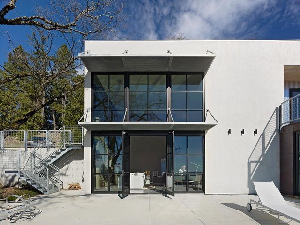 Exterior view of a contemporary two-storey white rendered building with a projecting flat roof canopy, extensive dark-framed floor-to-ceiling glazing, and open bi-fold or hinged glass doors at ground level. A concrete terrace, metal stair and balustrades, outdoor lounge chair, and mature trees surround the facade.