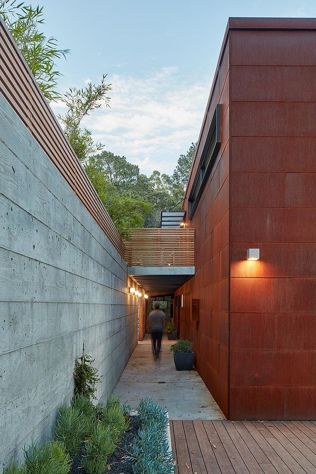 A narrow landscaped passage beside a modern building, with a long grey concrete wall on the left and a tall rust-coloured metal-clad facade on the right. Timber slatted screening and an overhead bridge-like connection span the walkway, which has concrete paving, timber decking, wall-mounted lighting, and drought-tolerant planting. The view leads toward a sheltered entry, with a person walking through the corridor.