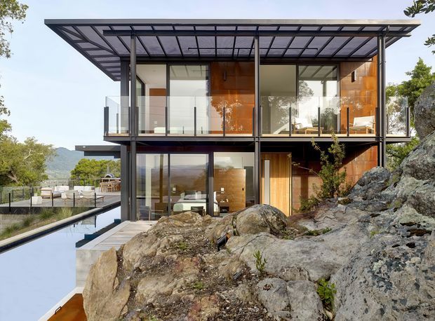 Contemporary multi-level residential house built into a rocky sloping site, with extensive floor-to-ceiling glazing, dark slender structural framing, warm timber wall cladding, a transparent flat roof canopy, glass balustrades, and a narrow reflecting pool or water feature beside an outdoor terrace. The home overlooks a wooded mountainous landscape.