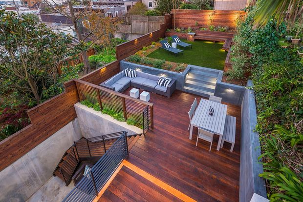 A multi-level landscaped residential outdoor entertaining area viewed from above. Dark timber decking forms stepped terraces with built-in grey seating, a dining table, lawn, timber privacy fencing, planted garden beds, and stairs with black metal balustrades. Warm integrated lighting highlights the deck edges and fences.