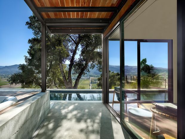 A contemporary covered outdoor terrace beside a narrow swimming pool, with a concrete bench or pool edge, timber-lined ceiling, dark metal framing, and large glazed sliding doors. The glazing reflects and reveals a warmly furnished interior sitting area. Beyond the terrace is a broad rural valley and mountain landscape, while mature trees provide shade.