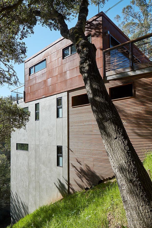 Exterior view of a contemporary multi-level home built into a sloping, wooded site. The facade combines rust-toned metal cladding, smooth grey concrete panels, and horizontal timber slats, with dark-framed windows and a projecting upper balcony screened by a metal framework. A mature tree dominates the foreground and partially obscures the building.