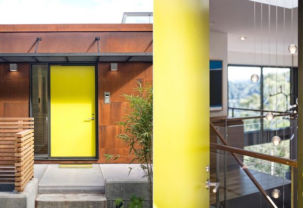 Split architectural view showing a bold yellow entry door set into a rust-toned metal-clad facade, alongside an interior stairwell with timber handrails, dark flooring, large glazing, and suspended pendant lights. The image highlights strong industrial materials and vivid colour contrast.