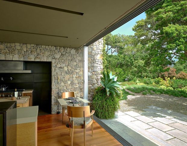 Open-plan kitchen and dining area with dark built-in cabinetry, pale stone island, timber flooring, and a compact dining table beside fully retracted floor-to-ceiling glazing. A rugged natural-stone wall continues outside into a landscaped courtyard with paving, gravel, and dense planting. The space has a strong indoor–outdoor connection and a refined, nature-integrated contemporary feel.