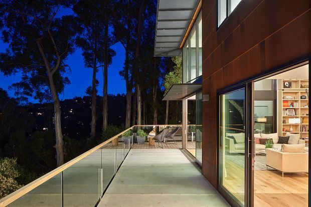 Twilight exterior view of a contemporary multi-level home with a broad timber-clad facade, extensive floor-to-ceiling glazing, and a large elevated terrace. The terrace has frameless glass balustrades, outdoor seating, and views into a warmly lit living room through open sliding doors. Tall trees and a dark wooded setting surround the property.