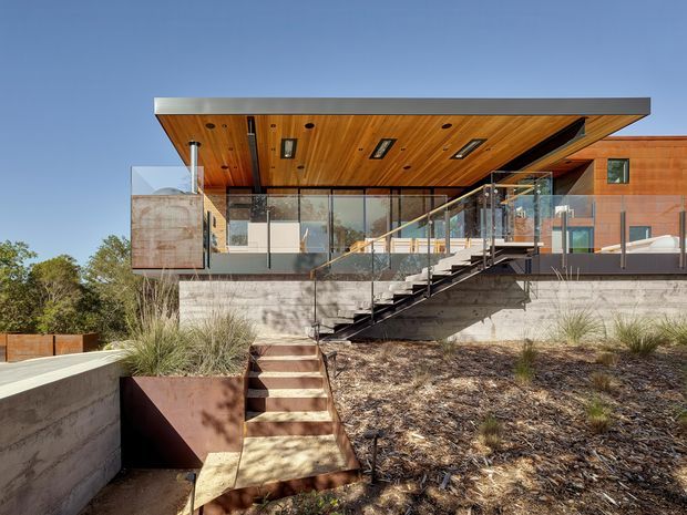 Exterior view of a contemporary elevated house set into a landscaped sloping site. The home features warm timber soffit lining, extensive glass balustrades and glazing, a broad flat roof with recessed skylights, exposed concrete foundations and stairs, and rust-toned metal-clad wall elements. Native grasses and dry ground cover surround the entry stair.
