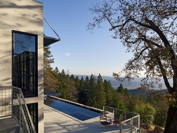Exterior architectural view of a modern multi-level residence on a wooded, elevated site. A tall pale rendered facade with dark floor-to-ceiling glazing and projecting balconies overlooks a narrow reflecting pool or swimming pool and outdoor terrace. Metal mesh balustrades, outdoor chairs, mature trees, and distant hills are visible under clear daylight.