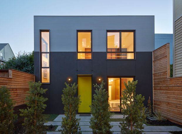 Front exterior view of a compact two-storey contemporary home at dusk. The facade combines dark smooth panels on the lower level with horizontal blue-grey cladding above, tall narrow and square windows glowing warmly from within, and a vivid yellow-green entry door. Timber privacy fencing, a paved stepping-stone path, and structured planting frame the frontage.