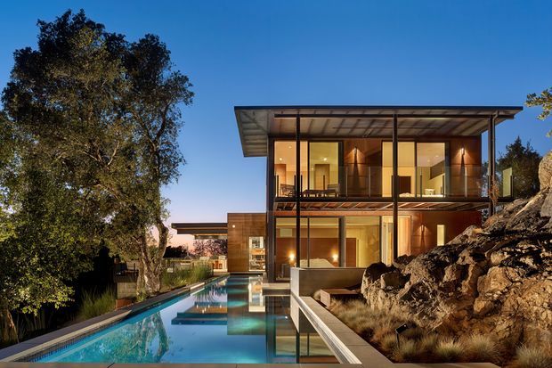 A modern two-storey glass-fronted residence set into a rocky, landscaped site, photographed at dusk. The building has a broad flat roof, extensive floor-to-ceiling glazing, warm timber-toned interior finishes, and a long reflective swimming pool extending through the foreground. Mature trees and natural rock formations frame the house.