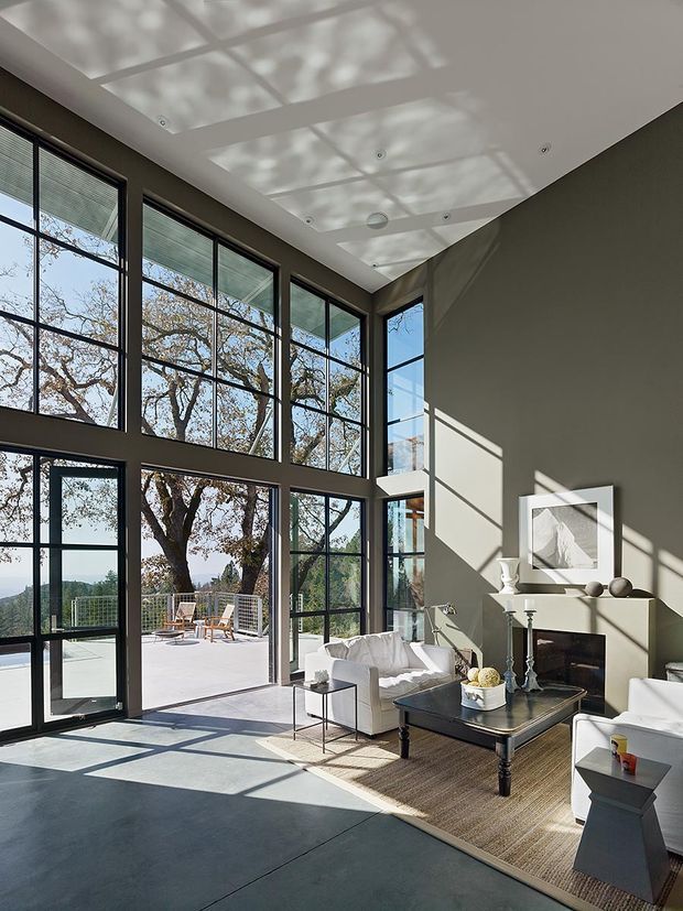 Double-height contemporary living room with expansive black-framed floor-to-ceiling glazing, a white ceiling, dark grey walls, and polished concrete flooring. White sofas and armchairs surround a dark coffee table and fireplace, while strong sunlight creates geometric shadows across the room. The glazing opens to an outdoor terrace with landscape views.