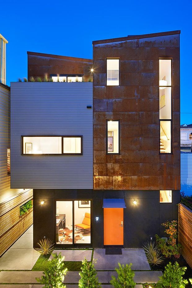 A tall, narrow contemporary urban residence photographed at dusk. The multi-storey facade combines weathered rust-toned metal cladding, grey horizontal siding, dark lower-level walls, and narrow vertical windows with warm interior illumination. An orange entry door sits beneath a small blue awning, opening onto a landscaped front path with concrete slabs, timber fencing, and drought-tolerant planting. The composition emphasizes the building exterior and its layered modern facade.