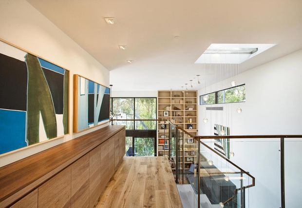 A bright contemporary upper-level landing or hallway overlooking a double-height living space. The scene features a long built-in timber storage cabinet beneath large abstract artworks, wide-plank timber flooring, a full-height glazed opening, a timber open bookshelf, a skylight, and a glass-and-dark-wood balustrade around the stairwell. White walls and ceilings create an airy, refined interior with strong natural light.