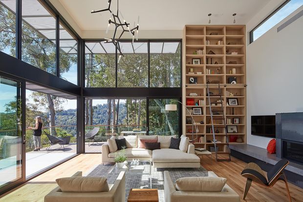 A bright, spacious contemporary living room with floor-to-ceiling black-framed glazing opening to an outdoor terrace and leafy views. The room features a tall built-in timber shelving wall with a ladder, a large cream sectional sofa, lounge chairs, a low coffee table, a wall-mounted television, and sculptural modern pendant lighting. Timber flooring and abundant natural light create an airy, refined atmosphere.