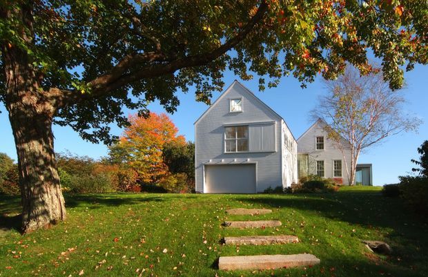Exterior view of a light grey, shingle-clad multi-storey house with a steep gabled roof, dormer window, projecting upper-level bay, and integrated garage. A stone stepping path crosses a broad lawn beneath mature trees, with autumn foliage surrounding the property.