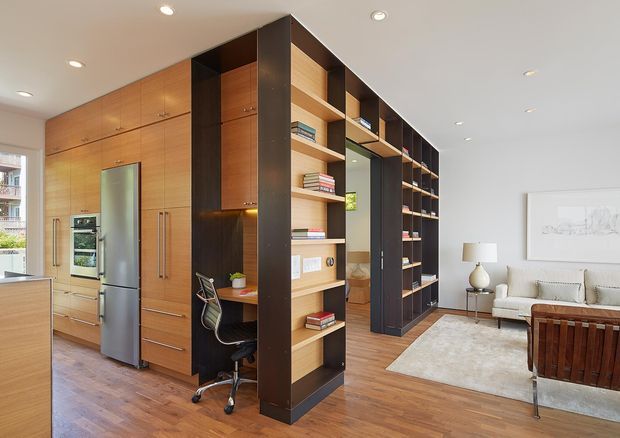 Open-plan contemporary interior featuring a warm timber kitchen with integrated stainless-steel appliances and a large custom built-in timber and dark-wood storage wall. The joinery incorporates open bookshelves, a compact home office nook, and a passage toward a bright living area with a light sofa, rug, side table, and artwork. Recessed ceiling lights and timber flooring create a cohesive, refined space.