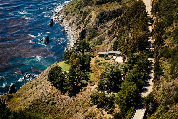 Aerial view of a rugged coastal property set on a steep, densely vegetated cliff above rocky blue ocean waters. A small low-rise building with a dark pitched roof sits among mature trees and landscaped lawn, while a narrow winding road runs along the hillside. The scene is natural, secluded, and distinctly coastal.