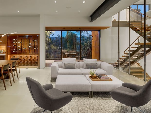Open-plan contemporary living room with a large light sectional sofa, ottoman, and two dark lounge chairs arranged on a textured rug. The room features a polished pale floor, expansive floor-to-ceiling glazing, exposed dark structural beam, timber-framed windows and doors, and a glass-sided staircase with timber treads. A timber-lined dining and wine display area is visible to the left, while bushland can be seen beyond the windows.
