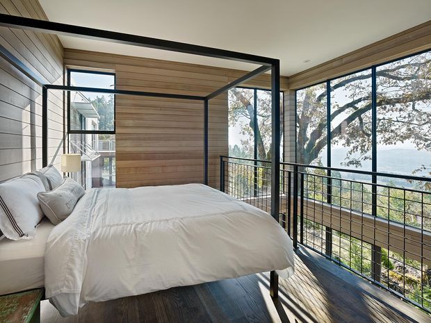 A bright modern bedroom with a four-poster bed, white bedding, extensive floor-to-ceiling glazing, and a black metal balustrade overlooking a leafy landscape and water beyond. Warm horizontal timber wall panelling and dark timber flooring create a natural, contemporary retreat.