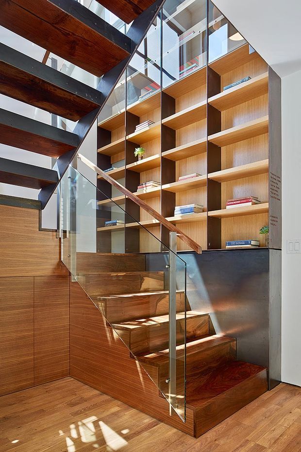Interior view of a contemporary stairway with rich timber treads and wall panelling, a clear glass balustrade, dark metal stringers, and a tall built-in timber bookshelf extending through multiple levels. Bright daylight enters through the glazed stairwell and highlights the warm wood surfaces.
