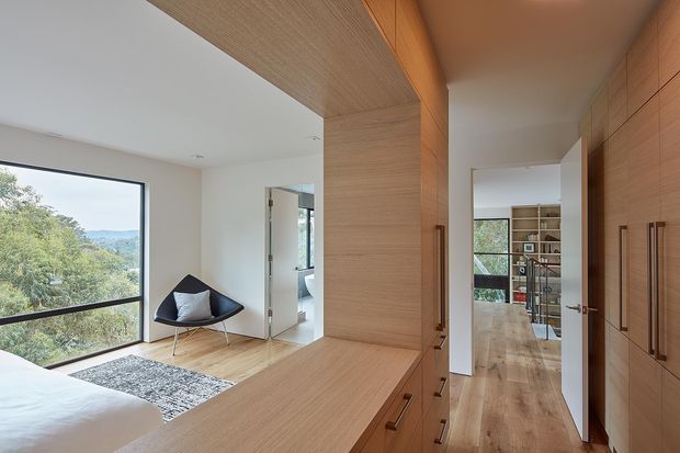 A bright contemporary bedroom with a large corner window overlooking greenery and distant hills. The room features pale timber flooring, extensive built-in timber cabinetry and joinery, a built-in desk or dresser, a black lounge chair with a cushion, and a grey rug. An open doorway leads toward a timber-floored circulation area and additional shelving.