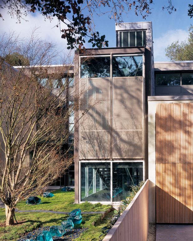 Exterior view of a contemporary multi-level residence with a tall glazed central volume, dark metal framing, translucent or textured facade panels, and warm vertical timber cladding. The building is set within a landscaped garden with mature trees, lawn, stone edging, and a narrow water feature or decorative garden installation. Strong geometric composition, extensive glazing, and an urban residential character are visible.