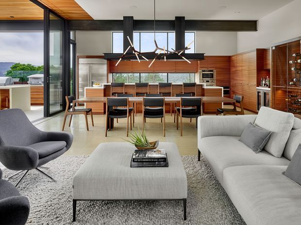 Open-plan contemporary living, dining and kitchen interior with floor-to-ceiling glazing, dark structural beams, warm timber ceiling and cabinetry, and a large dining table separating the kitchen from a light upholstered lounge area. A sculptural linear pendant hangs above the dining table, while a monochromatic seating arrangement and textured rug create a refined, comfortable setting. The glazing opens toward an outdoor terrace and landscape.
