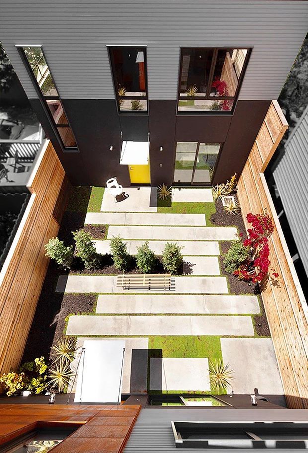 A high-angle view down into a narrow, enclosed landscaped courtyard between modern multi-storey buildings. The courtyard features staggered rectangular concrete stepping slabs set into lawn, dark gravel garden beds, small shrubs and ornamental grasses, timber privacy fencing, and several large windows and doors opening onto the space. Contemporary dark cladding and horizontal metal siding frame the courtyard, with a simple white outdoor chair and a linear bench visible.