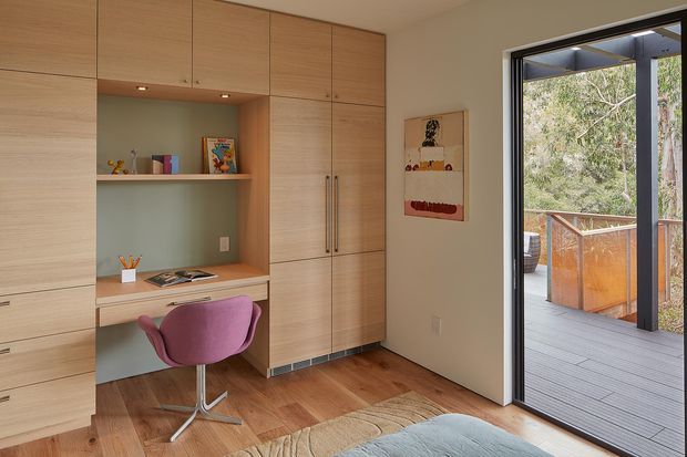 Compact, warm home office nook integrated into a full-height timber built-in joinery wall. The joinery includes overhead and side cupboards, open shelving, a desk surface, drawers, and a central workspace with a mauve swivel chair. Timber flooring and a partially visible rug add warmth, while a large glazed door opens to a timber outdoor deck and railing.