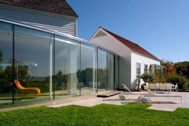 Exterior view of a traditional white weatherboard house with steep red-tiled pitched roofs and a modern glass-enclosed extension. The extension opens onto a paved terrace with outdoor dining furniture, while lawn and landscaped planting surround the building. Strong contrast between the older pitched-roof form and the transparent contemporary addition.