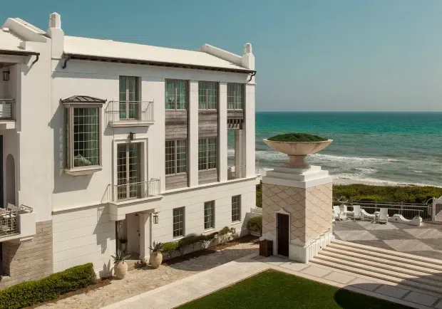 Exterior view of an elegant multi-storey coastal residence overlooking the ocean. The white rendered facade has tall multi-pane windows, projecting balconies with metal balustrades, classical trim, and a low pale roof with chimney elements. A landscaped courtyard includes clipped hedges, lawn, paved terraces, broad steps, white outdoor seating, and a decorative stone-clad pedestal with an urn planter.