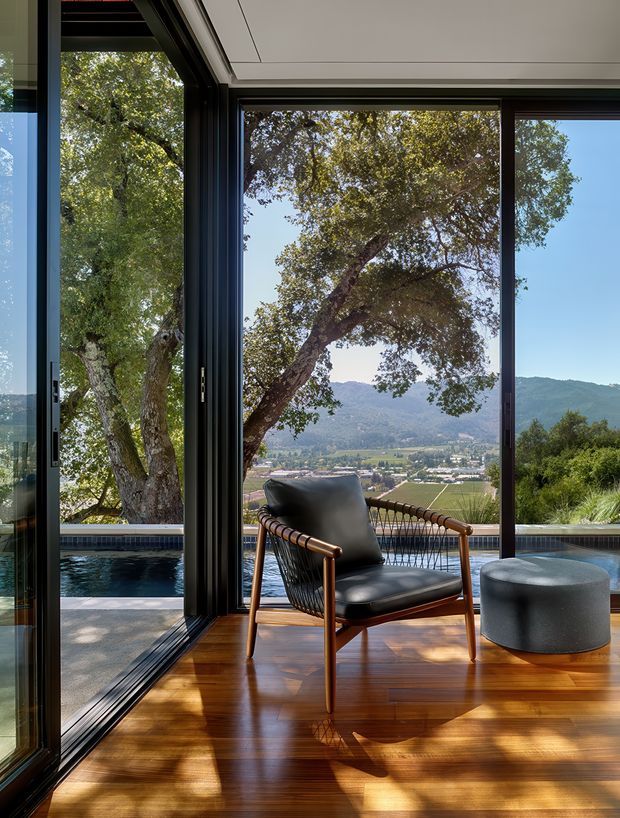 A refined indoor-outdoor living space with expansive black-framed floor-to-ceiling glazing overlooking a mountain valley and vineyard-like landscape. A mid-century-inspired leather lounge chair and round ottoman sit on a glossy timber floor beside a narrow reflecting or swimming pool. The room has a clean white ceiling with recessed detailing and strong visual connection to the landscape.