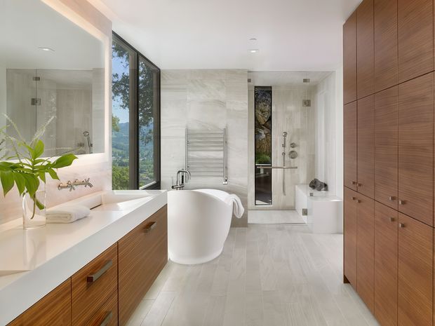 Bright, spacious contemporary bathroom with a freestanding white soaking tub, double vanity, large mirror, glass-enclosed shower, heated towel rail, and extensive warm timber cabinetry. Pale stone-look surfaces and floor tiles create a clean, refined atmosphere, while a tall window provides a leafy landscape view.