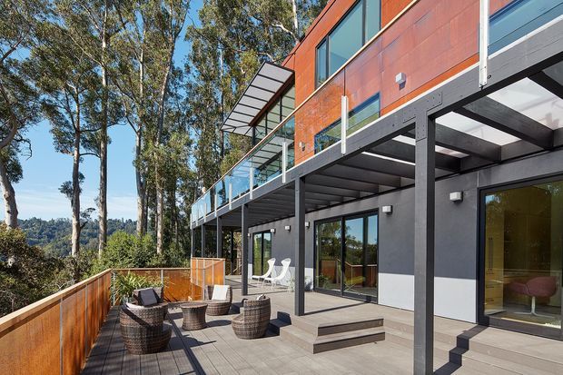 Exterior view of a contemporary multi-level house set among tall trees, with a large timber deck, glass balustrades, dark structural framing, extensive glazing, and a distinctive rust-red upper cladding volume. Outdoor seating and steps connect the terrace to the glazed ground level.