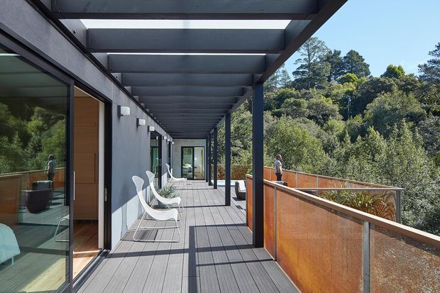 A long, covered exterior deck or balcony walkway alongside a modern house, with dark steel posts and beams forming a pergola-like roof structure. Full-height glazing and timber-framed doors open onto the deck, while perforated rust-toned metal balustrades overlook dense bushland. White outdoor lounge chairs and strong linear shadows add to the contemporary architectural character.