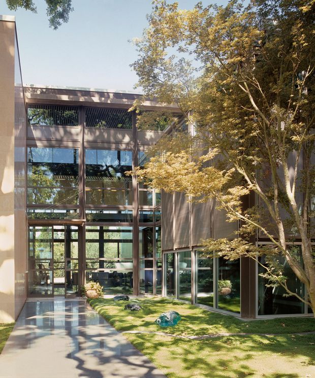 A contemporary multi-storey building arranged around a landscaped courtyard, viewed from an exterior walkway. The facade combines extensive floor-to-ceiling glazing, dark metal window frames, vertical structural elements, and warm brown cladding or screens. Mature trees, clipped lawn, sculptural rocks, and reflected sunlight soften the architectural composition. The building has a flat roofline and a strong indoor-outdoor relationship.