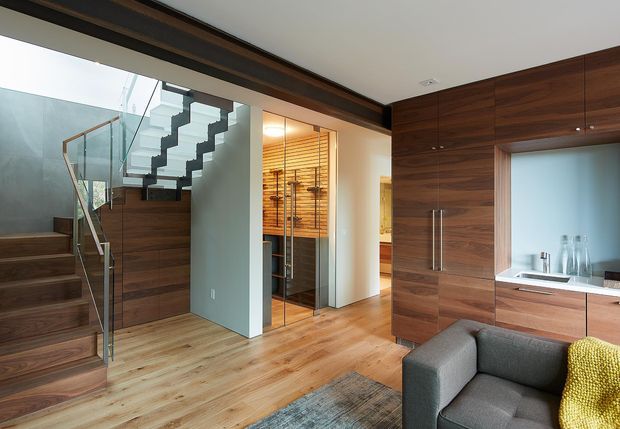 A contemporary open-plan interior featuring a timber staircase with glass balustrades, extensive dark timber wall panelling and cabinetry, light timber flooring, and a compact bar or display area beneath warm linear lighting. A grey sofa occupies the foreground beside a kitchen or kitchenette zone with a pale benchtop. The space is refined and warm, with strong horizontal timber detailing and clean modern lines.