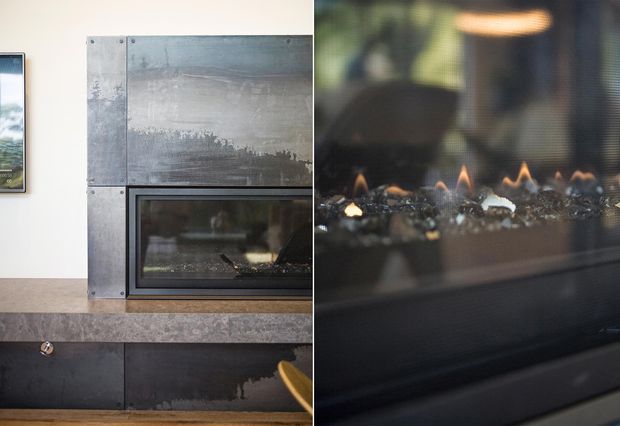 Close-up interior view of a contemporary built-in gas fireplace. The fireplace is framed with dark, weathered metal panels and a broad mottled stone or concrete hearth and mantel surface. A glass-front firebox with visible flames and decorative stones dominates the right side, while dark built-in cabinetry sits below and a framed artwork is partially visible at left.